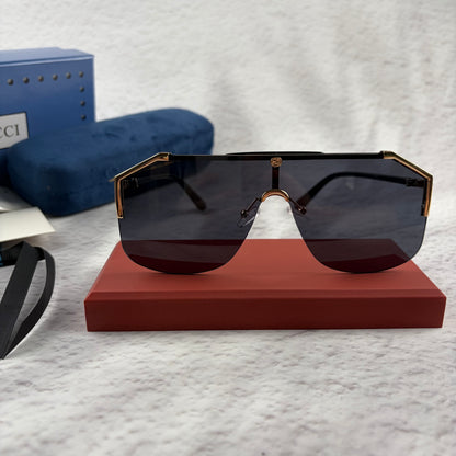 Oversized shield sunglasses with a gold metal frame and dark black lenses on a red display stand.