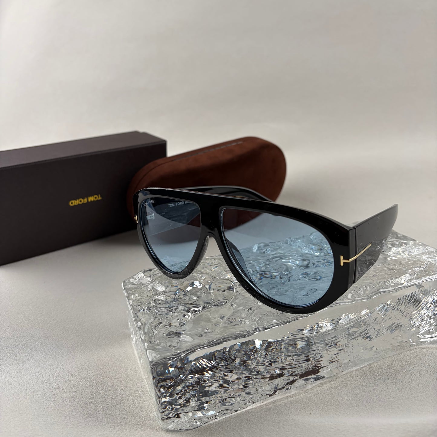 Black acetate oversized aviator sunglasses with blue tinted lenses resting on a clear ice-like block