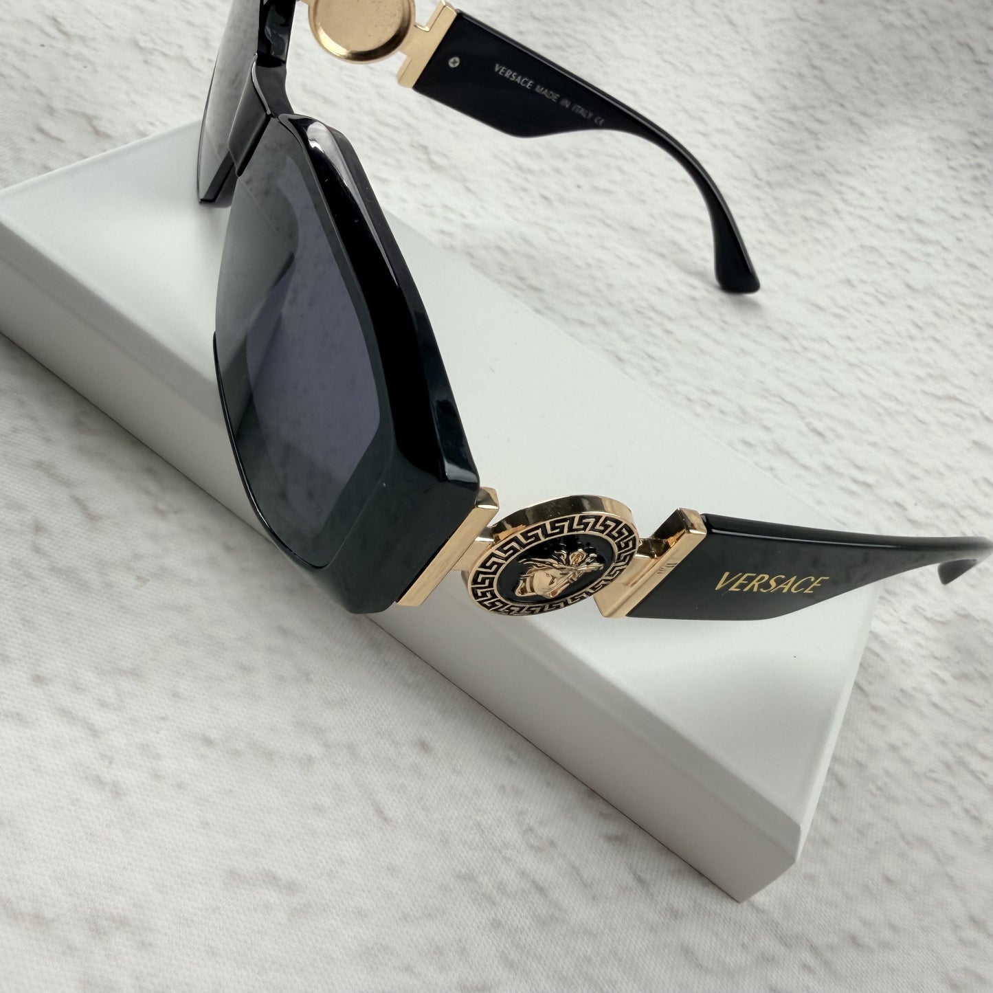 Black geometric sunglasses with gold side emblems resting on a white display block.
