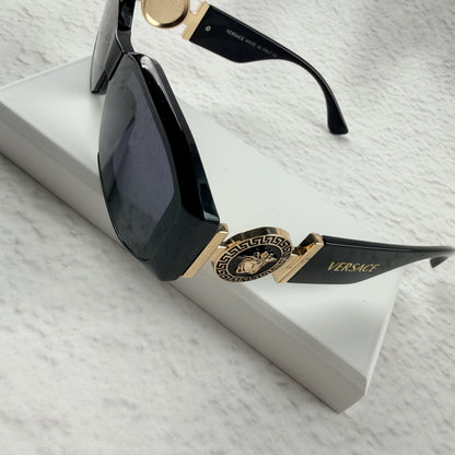 Black geometric sunglasses with gold side emblems resting on a white display block.