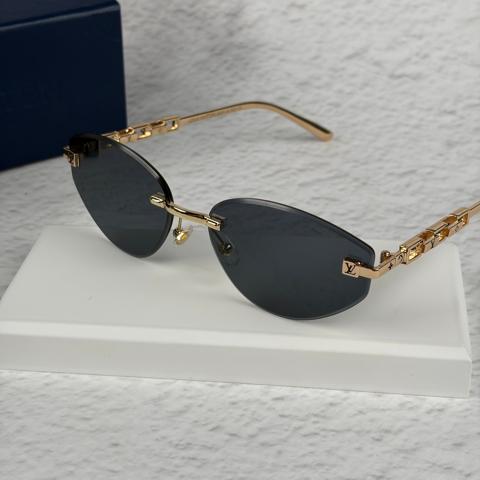 Rimless sunglasses with gold metal arms and dark gradient lenses resting on a white display block.
