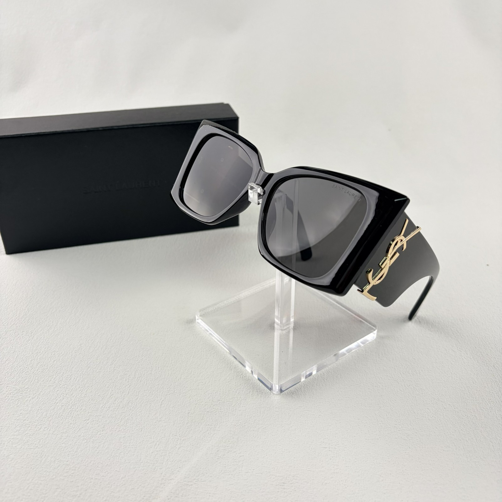Pair of oversized black square sunglasses with gold metal logo accents on temples on display stand.