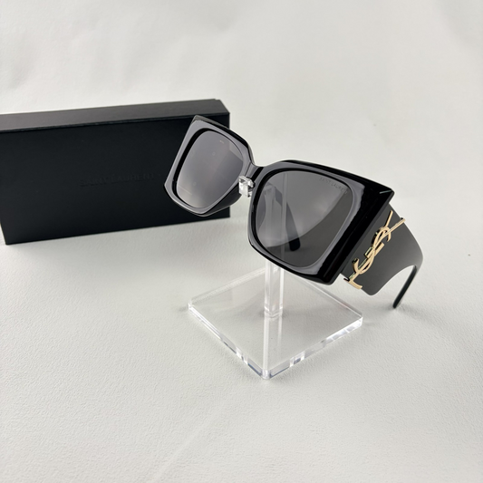 Pair of oversized black square sunglasses with gold metal logo accents on temples on display stand.