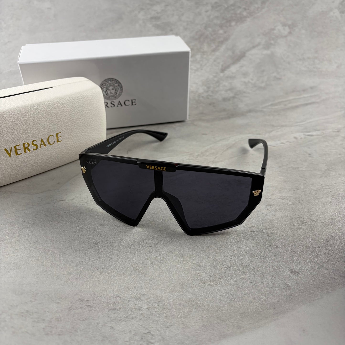 Oversized black shield sunglasses with gold metal accents on arms resting on a blue display box.
