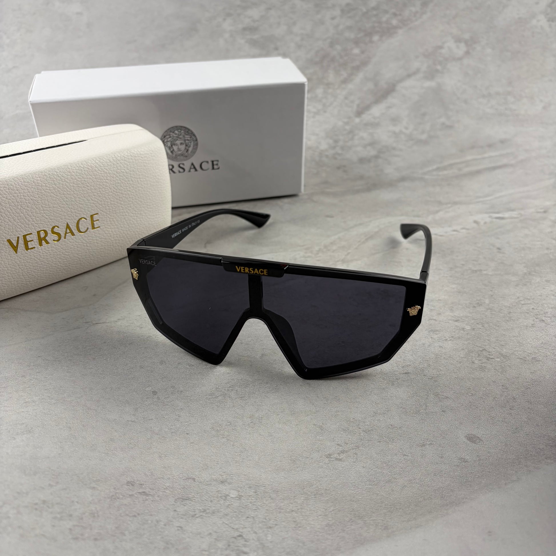 Oversized black shield sunglasses with gold metal accents on arms resting on a blue display box.