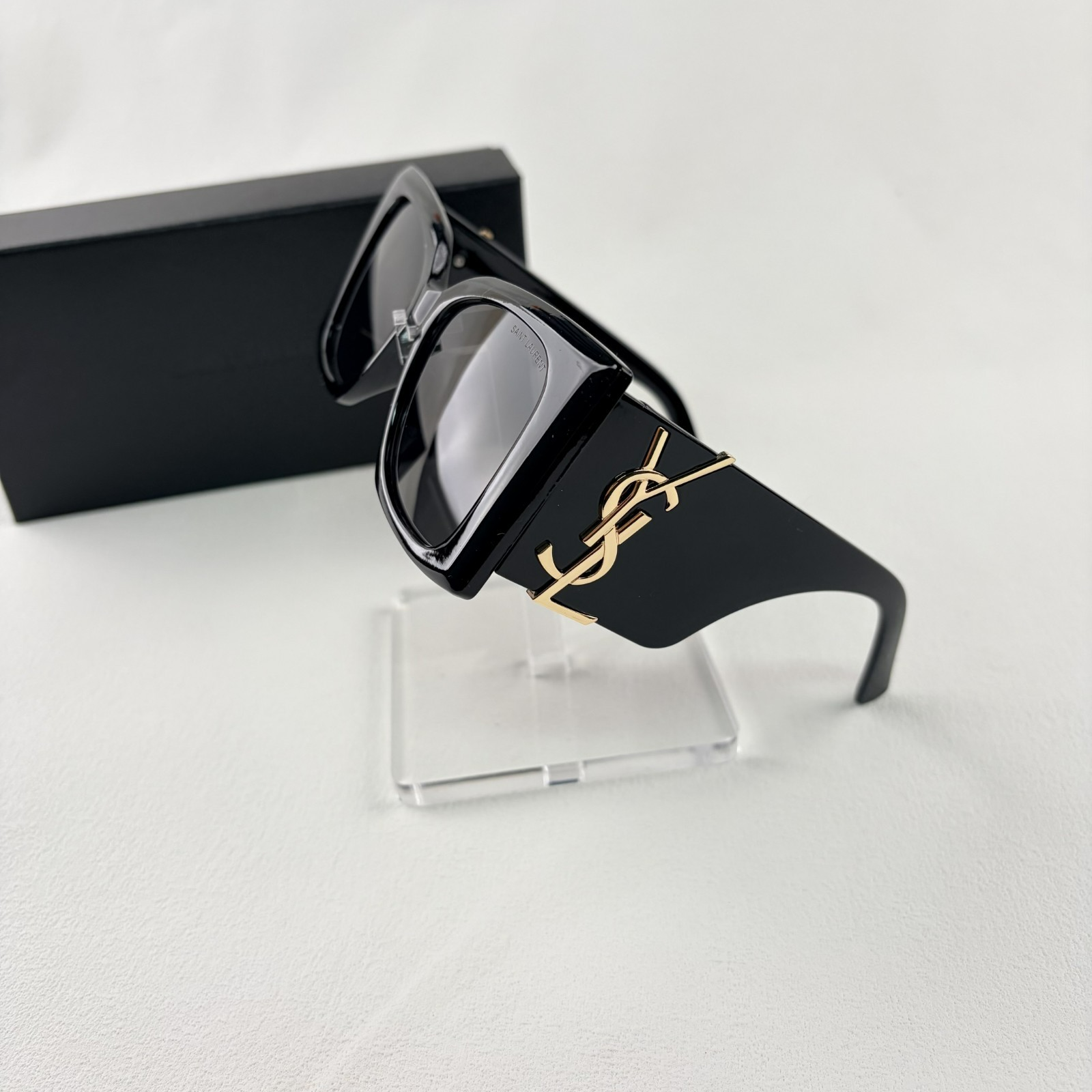 Pair of oversized black square sunglasses with gold metal logo accents on temples on display stand.