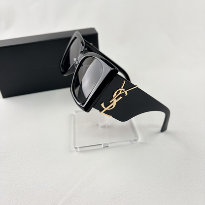 Pair of oversized black square sunglasses with gold metal logo accents on temples on display stand.