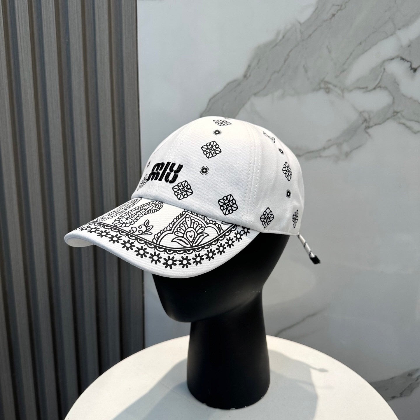 White cotton baseball cap with black paisley patterns and an adjustable drawstring back closure.
