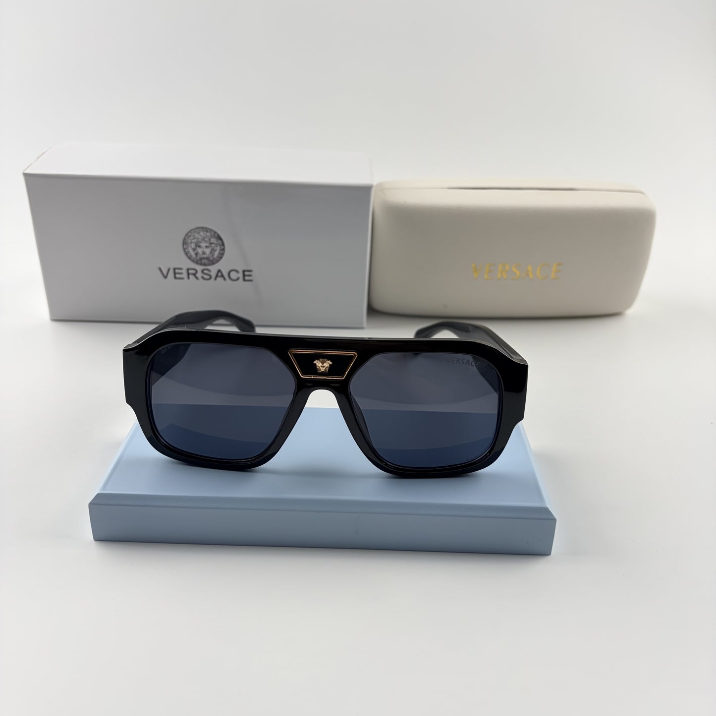 Black oversized aviator sunglasses with gold emblem on bridge, dark lenses, set on a blue stand.