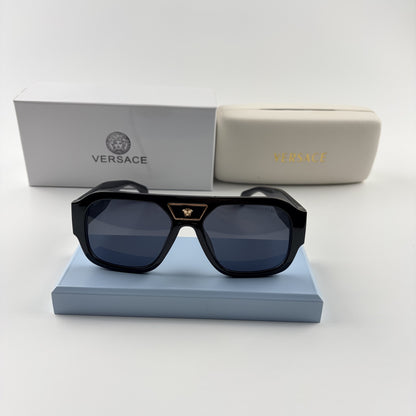 Black oversized aviator sunglasses with gold emblem on bridge, dark lenses, set on a blue stand.