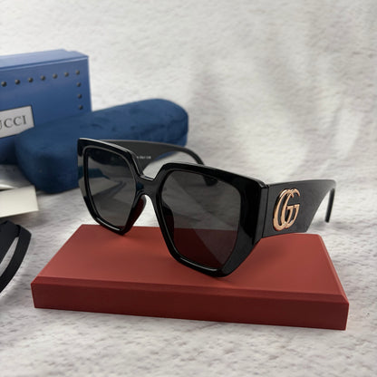 Oversized black square sunglasses with metallic temple detail, resting on a red display box.