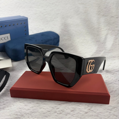 Oversized black square sunglasses with metallic temple detail, resting on a red display box.