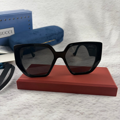 Oversized black square sunglasses with metallic temple detail, resting on a red display box.