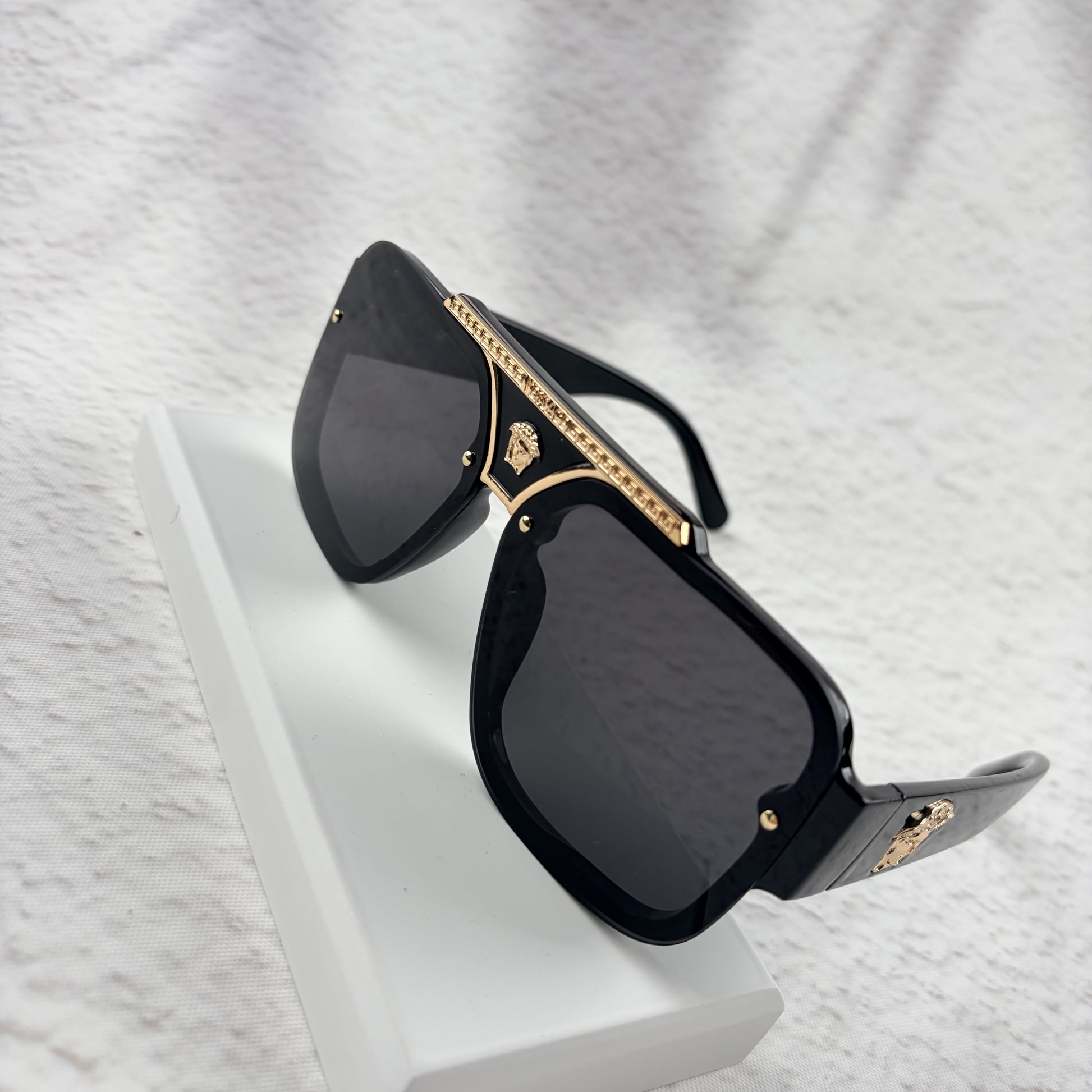 Oversized black flat-top sunglasses with a gold metal bridge and gold emblems on the temples.