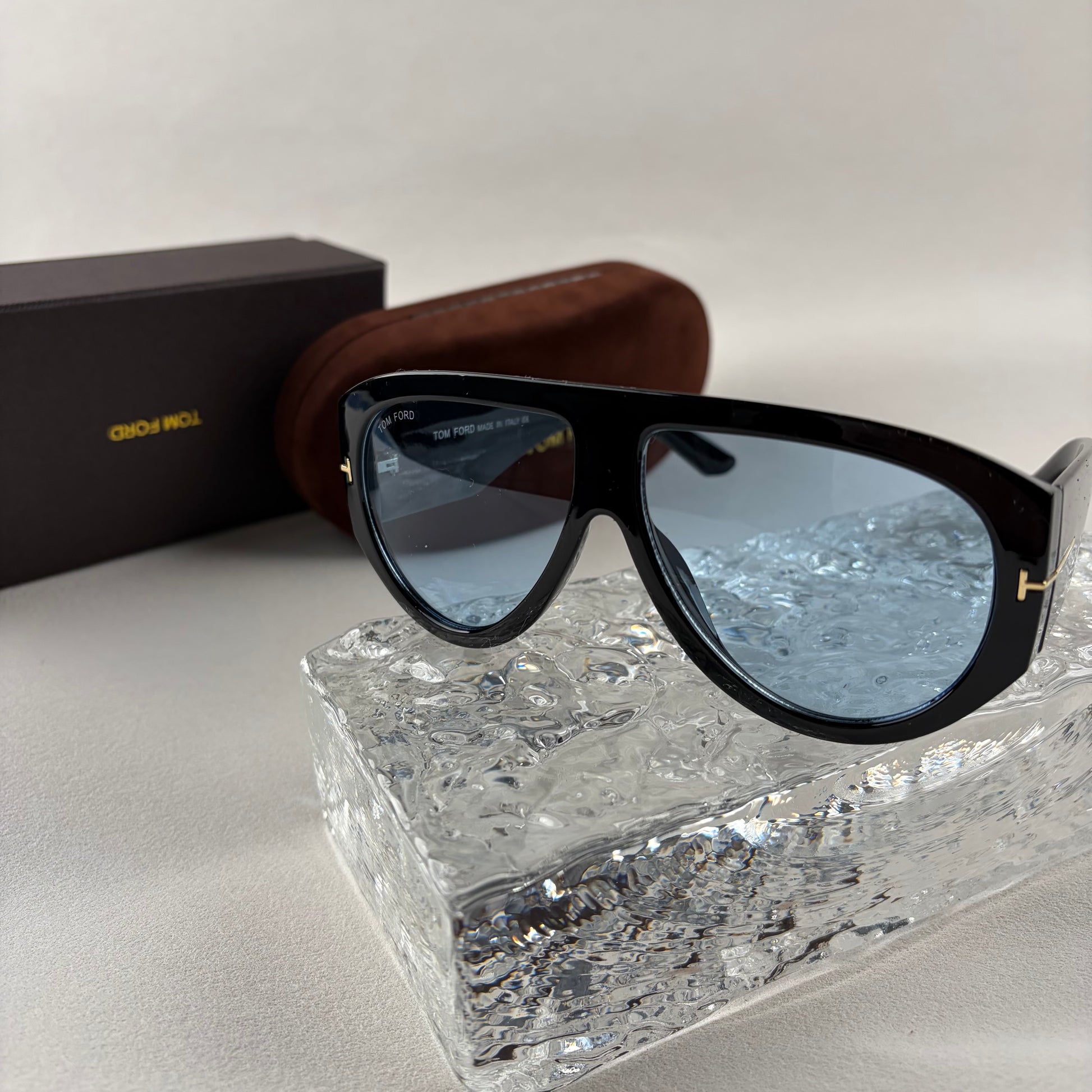 Black acetate oversized aviator sunglasses with blue tinted lenses resting on a clear ice-like block