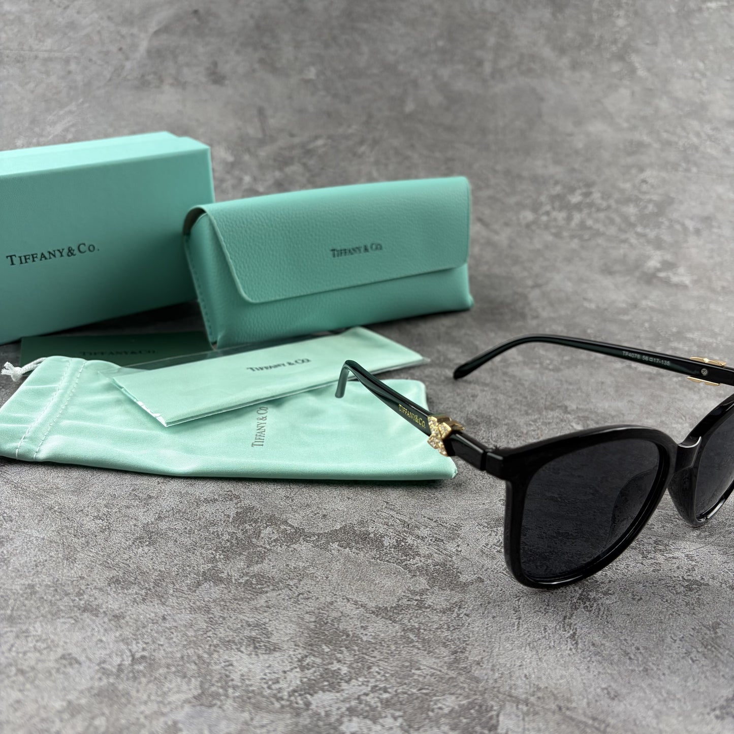 Black cat-eye sunglasses with decorative side temple accents, presented with a branded protective ca