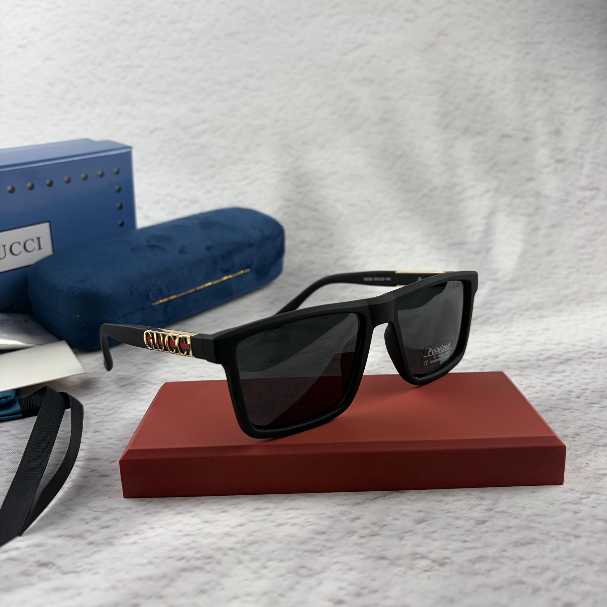 Matte black polarized sunglasses with gold logo detailing on temple arms, sitting on a red display b