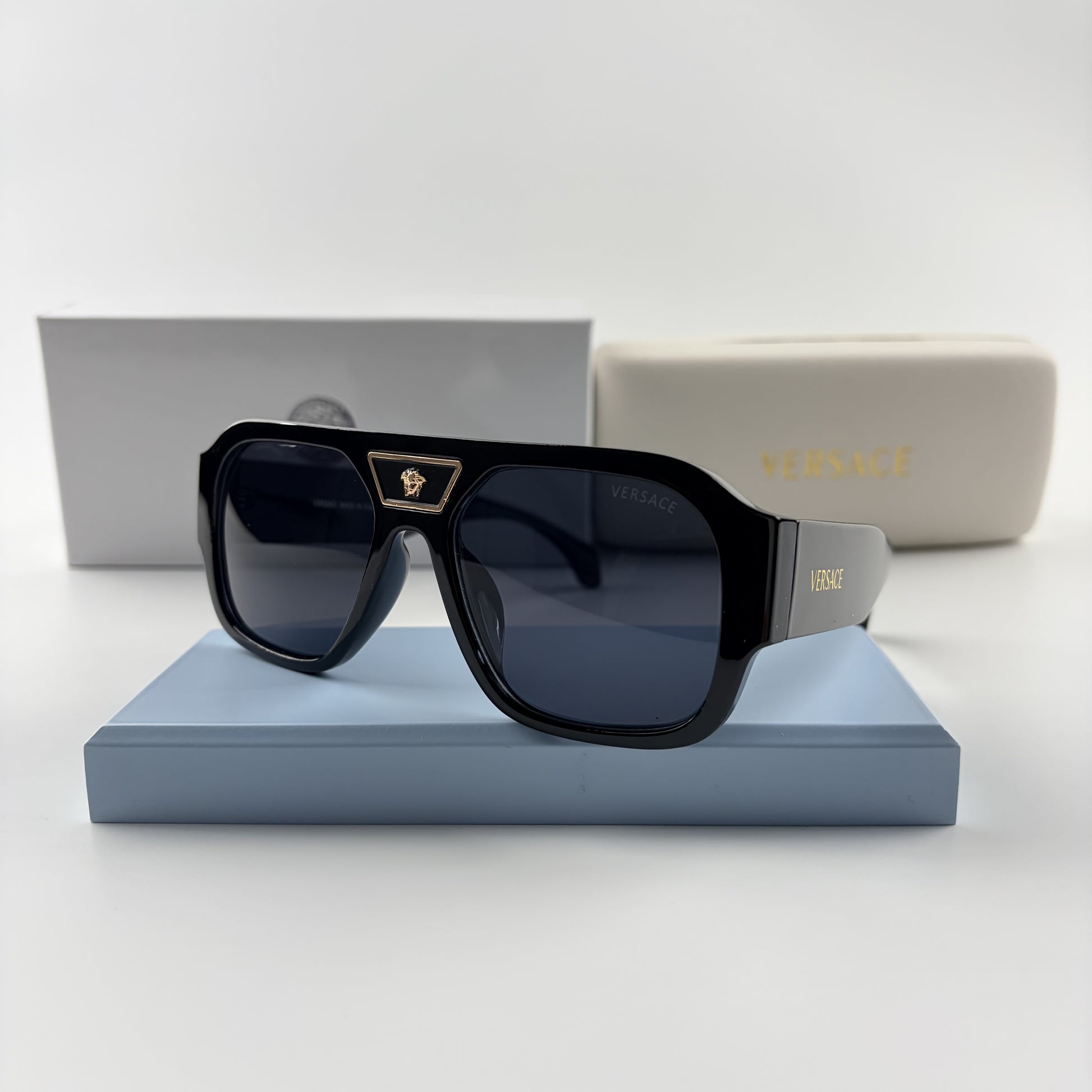 Black oversized aviator sunglasses with gold emblem on bridge, dark lenses, set on a blue stand.