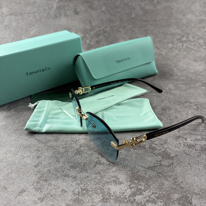 Rimless sunglasses with gradient blue lenses and gold-tone metal arms, displayed with protective cas