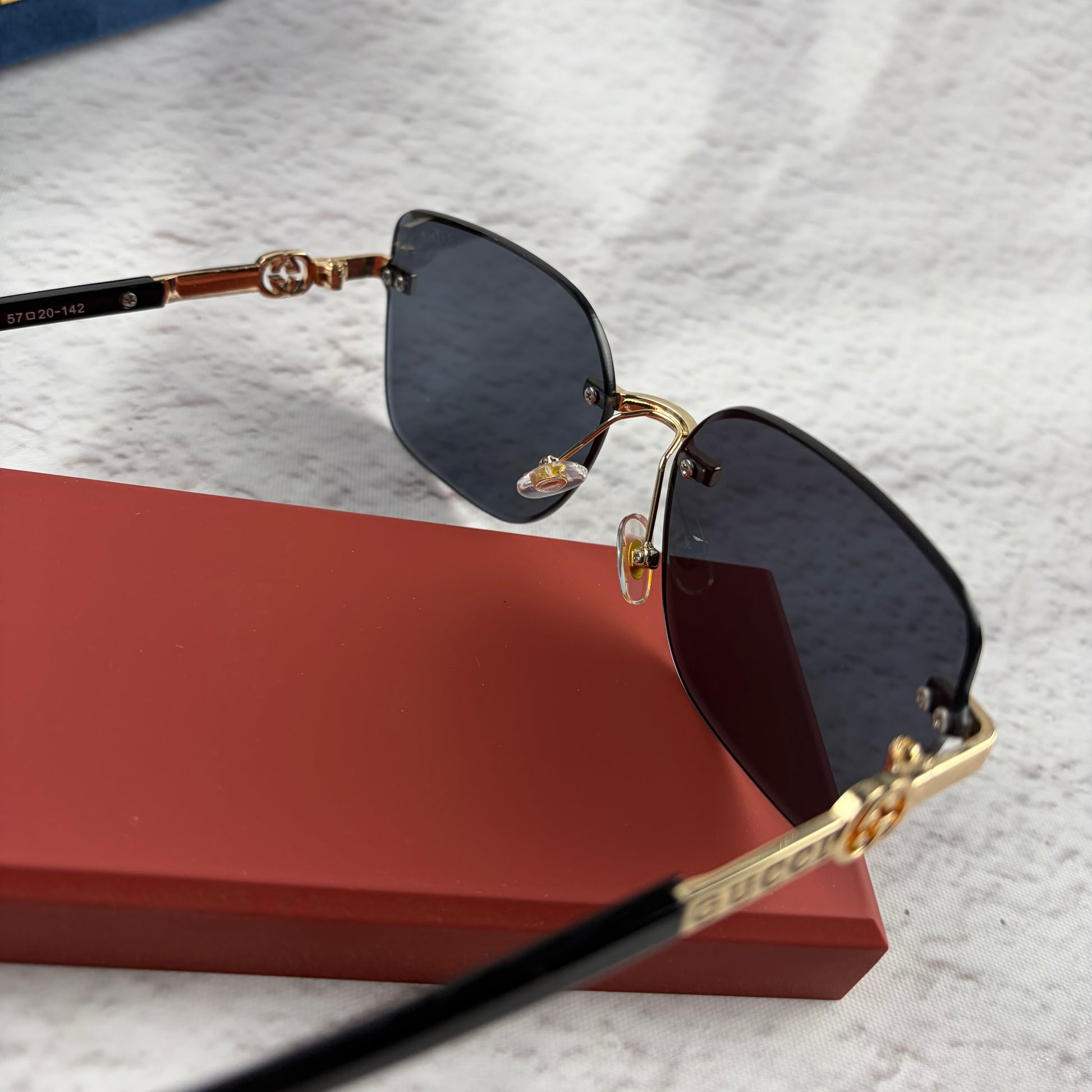 Rimless rectangular sunglasses with gold-tone metal arms and dark lenses on a red display stand.