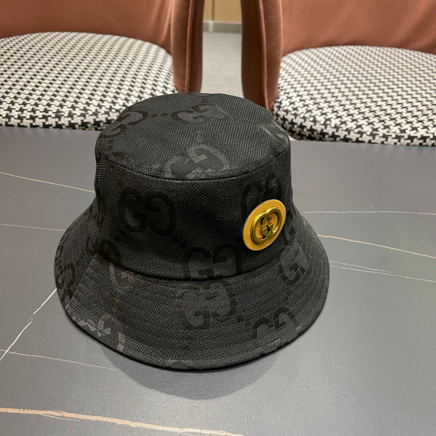 A black textured bucket hat with a circular gold emblem, featuring a monogrammed fabric design.