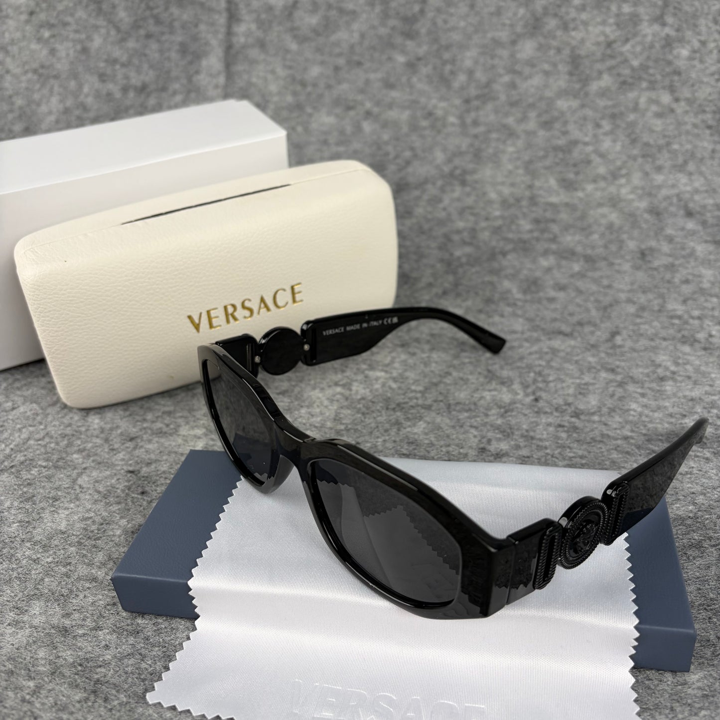 A pair of sleek black oval sunglasses resting on a cleaning cloth next to a white protective case.