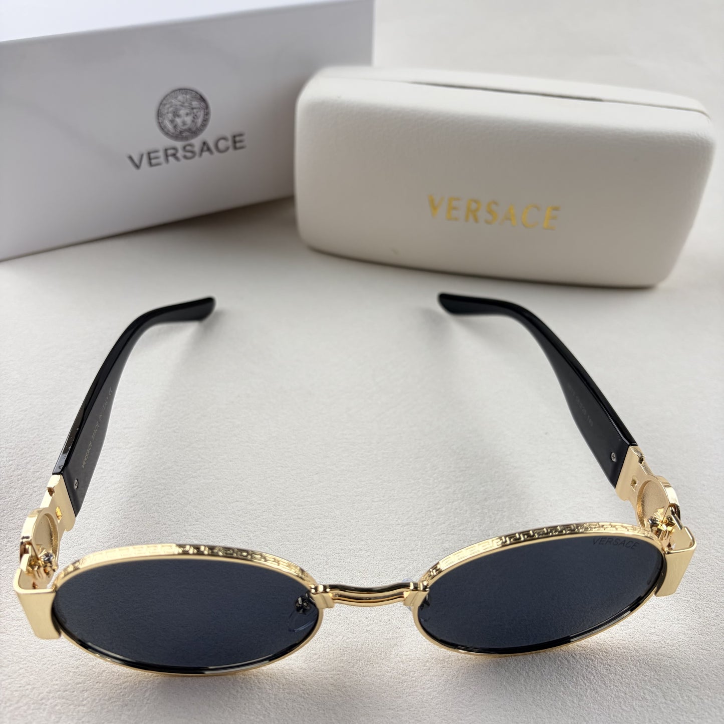 Oval gold metal frame sunglasses with black temples and dark tinted lenses, shown with a storage cas