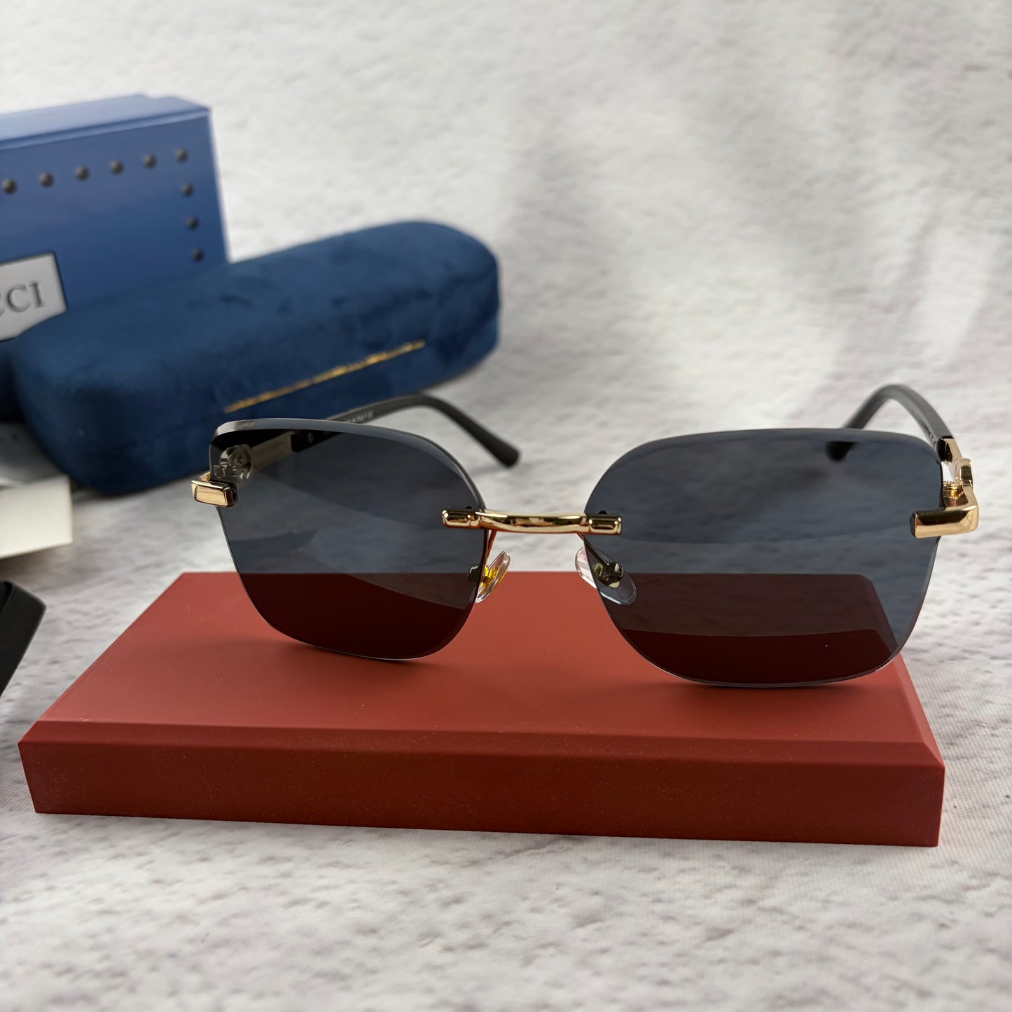 Rimless rectangular sunglasses with gold-tone metal arms and dark lenses on a red display stand.