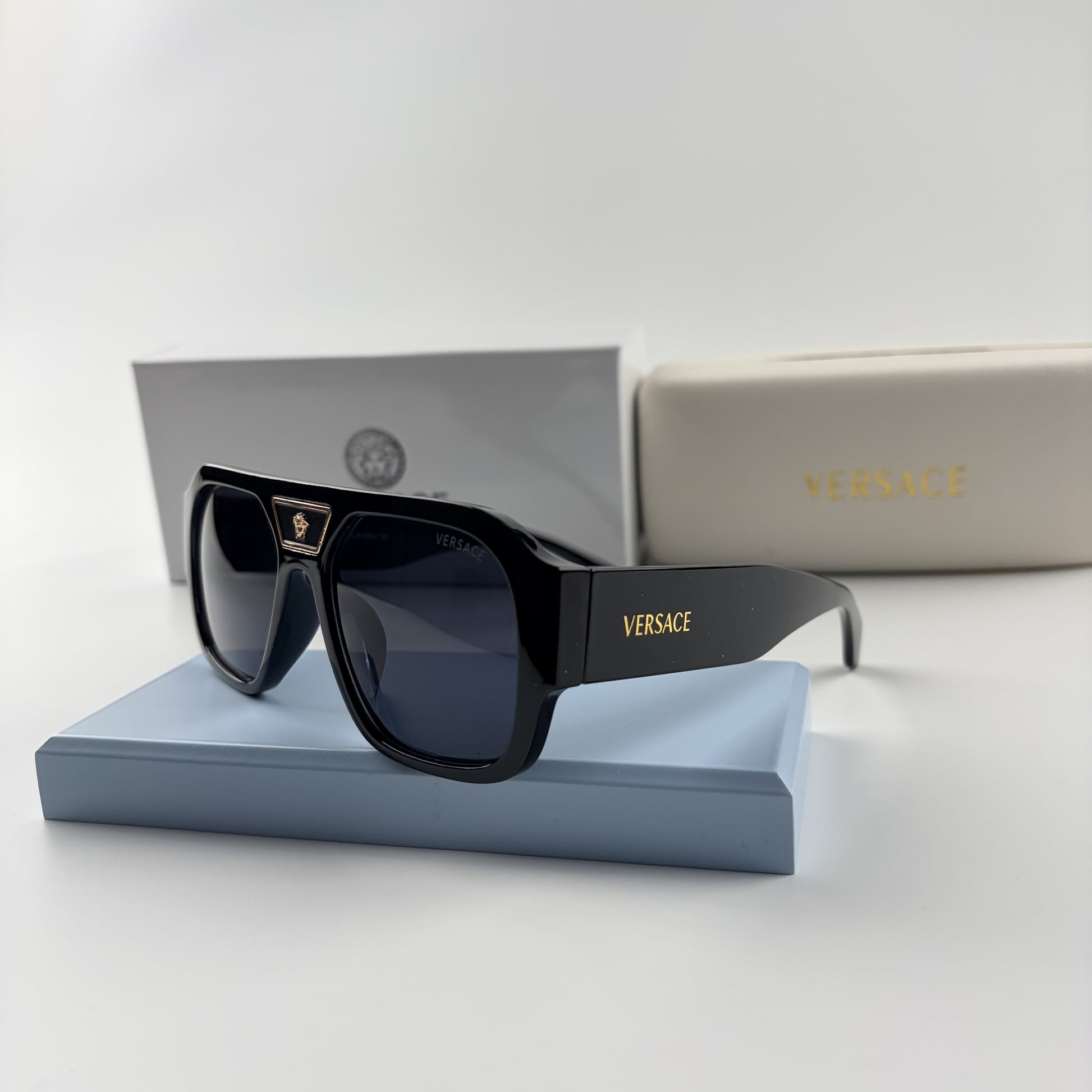 Black oversized aviator sunglasses with gold emblem on bridge, dark lenses, set on a blue stand.