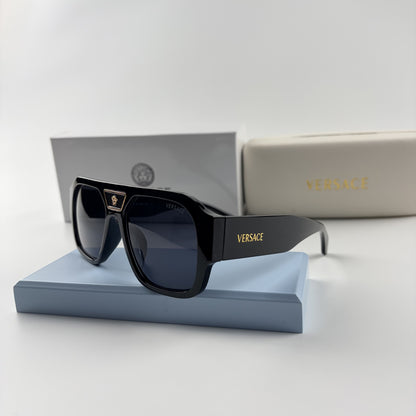 Black oversized aviator sunglasses with gold emblem on bridge, dark lenses, set on a blue stand.