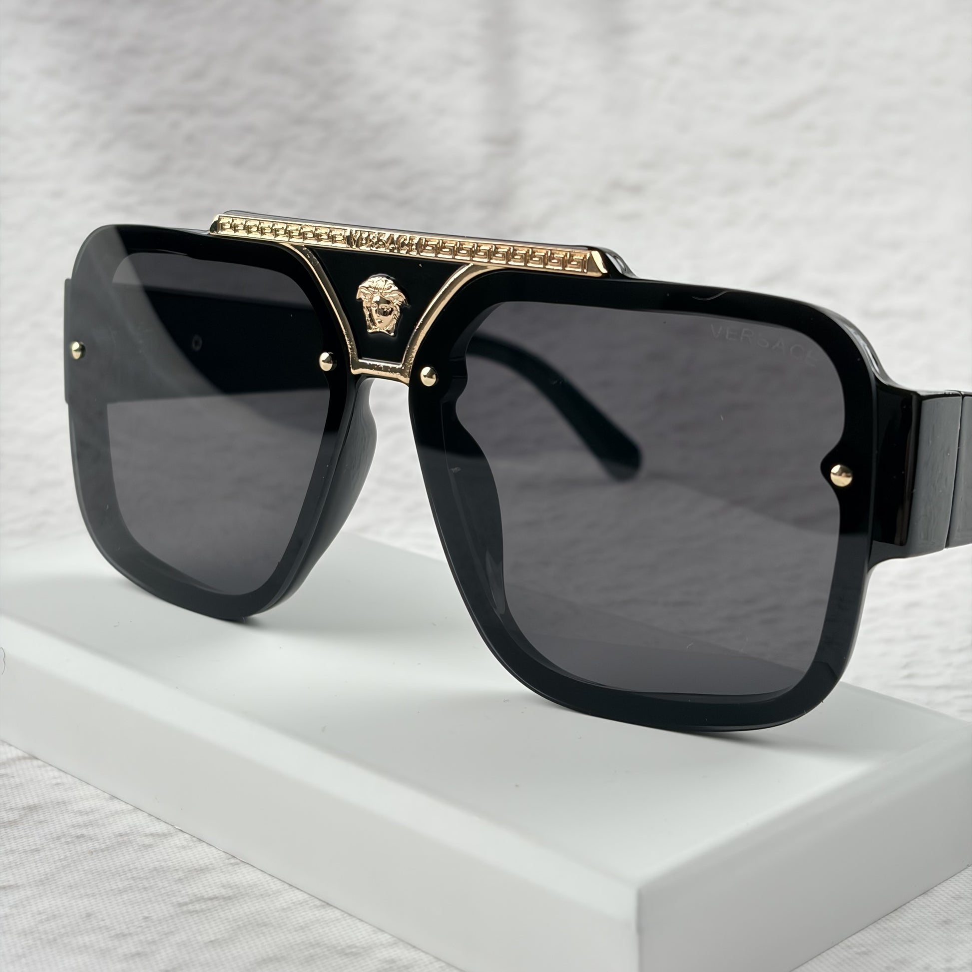 Oversized black flat-top sunglasses with a gold metal bridge and gold emblems on the temples.