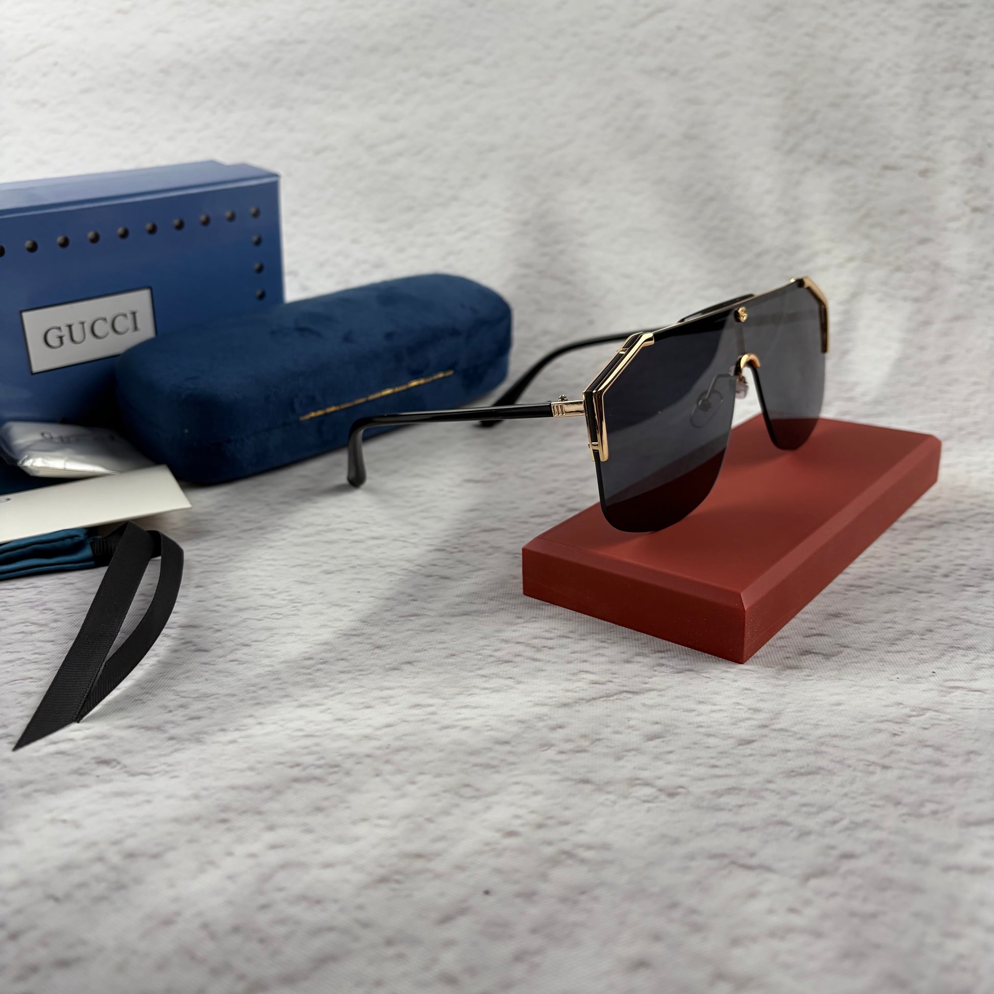 Oversized shield sunglasses with a gold metal frame and dark black lenses on a red display stand.