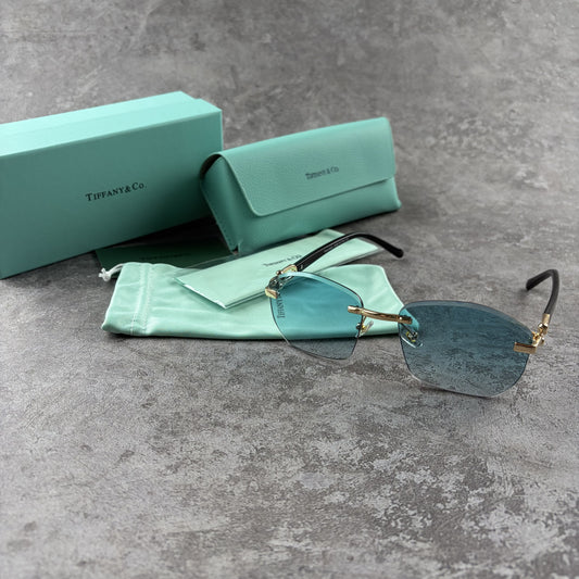 Rimless sunglasses with gradient blue lenses and gold-tone metal arms, displayed with protective cas