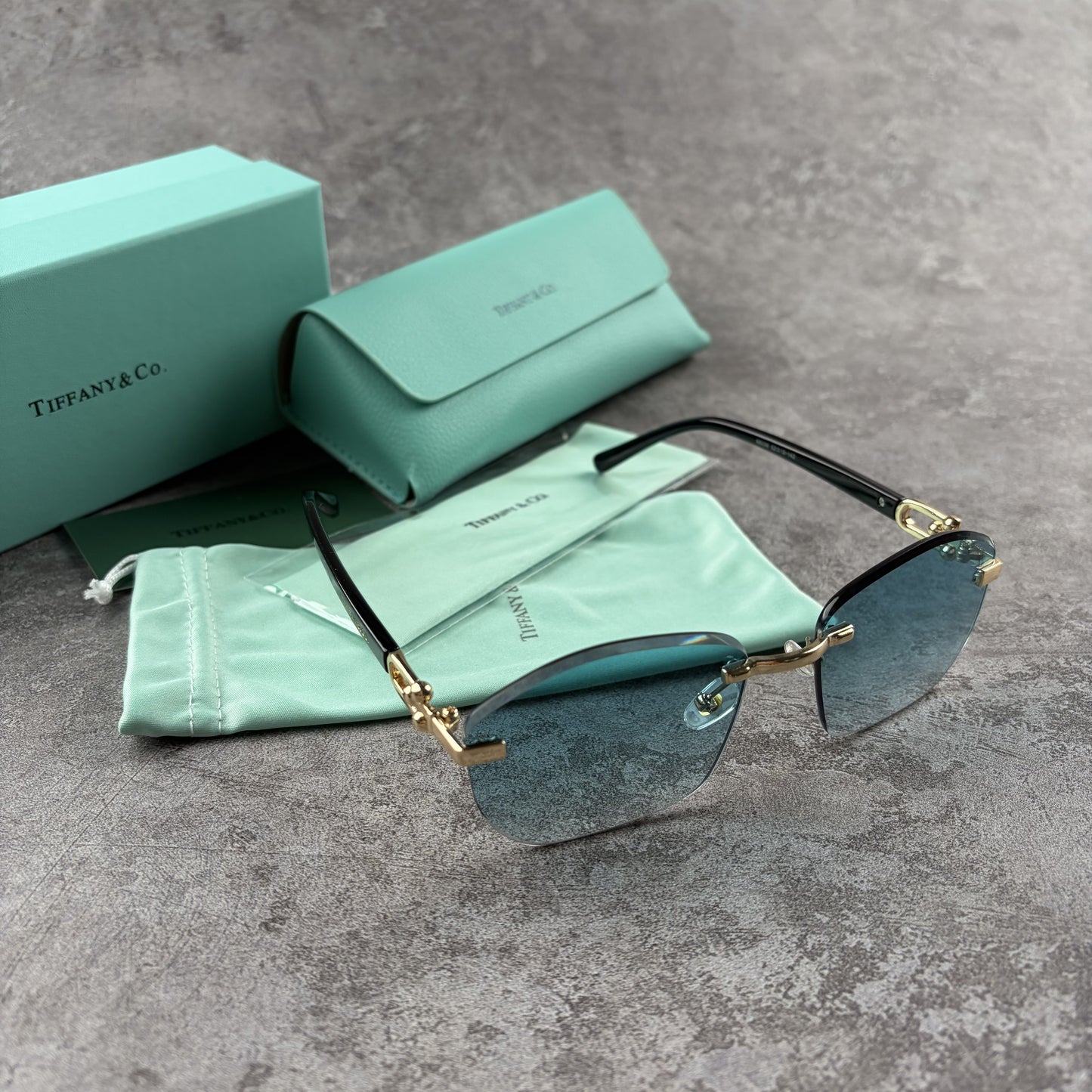 Rimless sunglasses with gradient blue lenses and gold-tone metal arms, displayed with protective cas