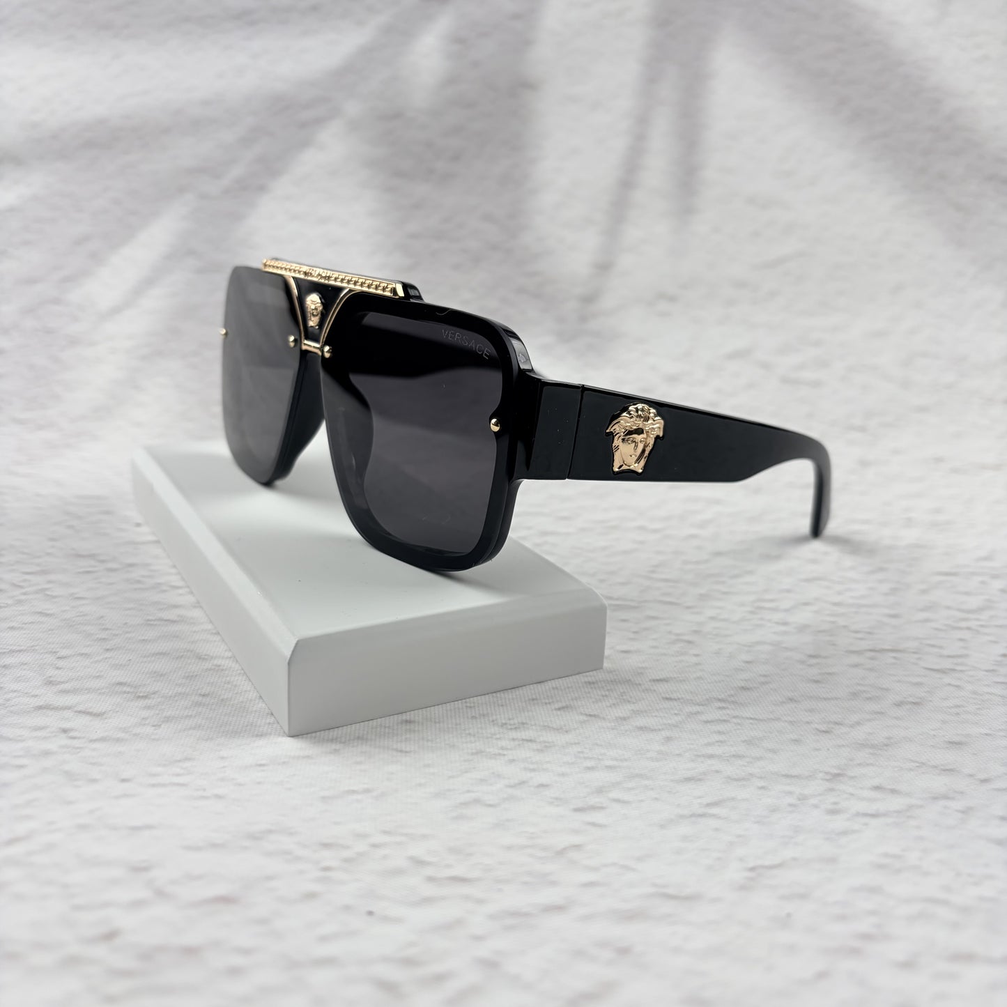 Oversized black flat-top sunglasses with a gold metal bridge and gold emblems on the temples.