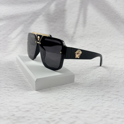 Oversized black flat-top sunglasses with a gold metal bridge and gold emblems on the temples.