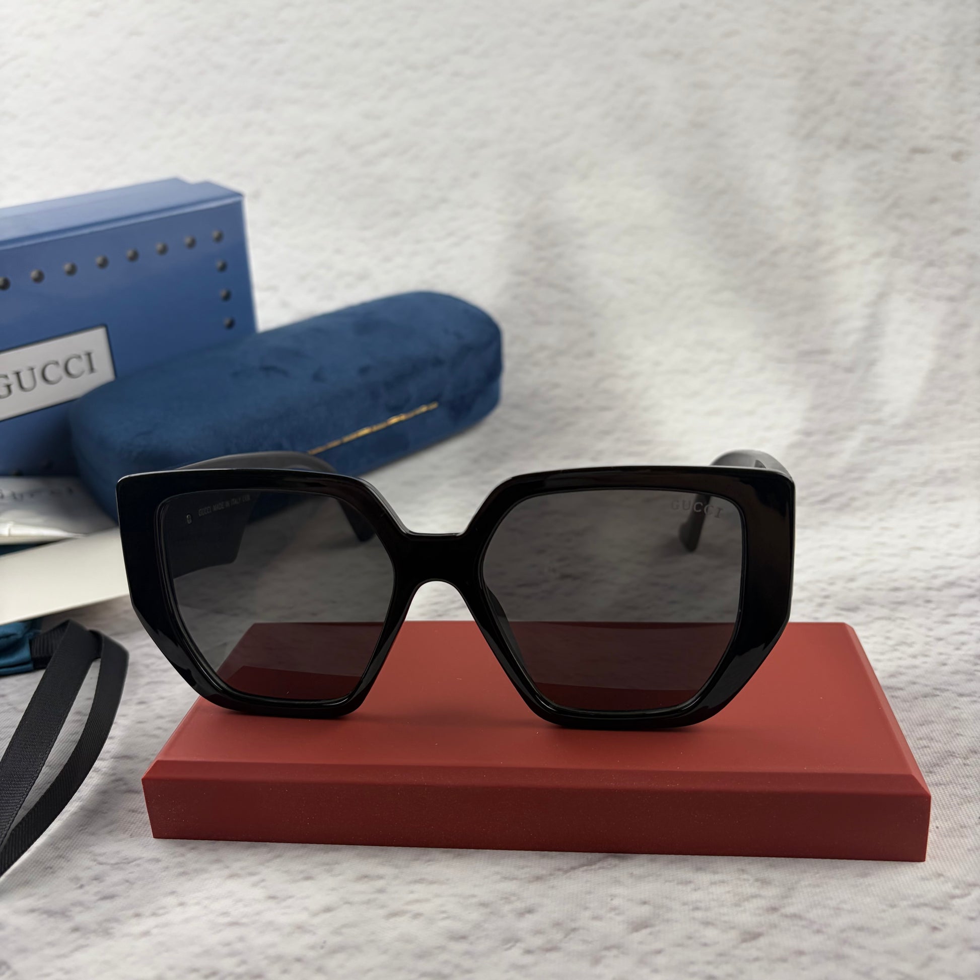 Oversized black square sunglasses with metallic temple detail, resting on a red display box.