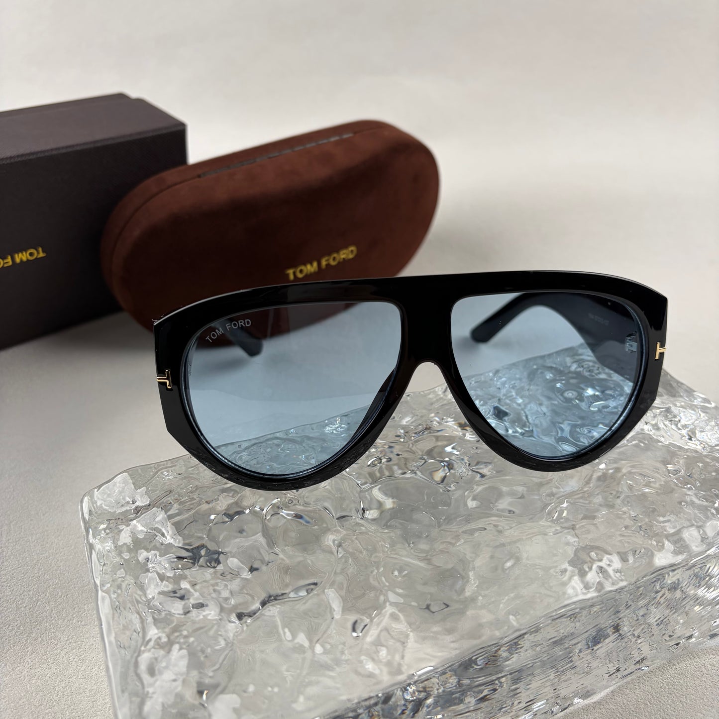 Black acetate oversized aviator sunglasses with blue tinted lenses resting on a clear ice-like block