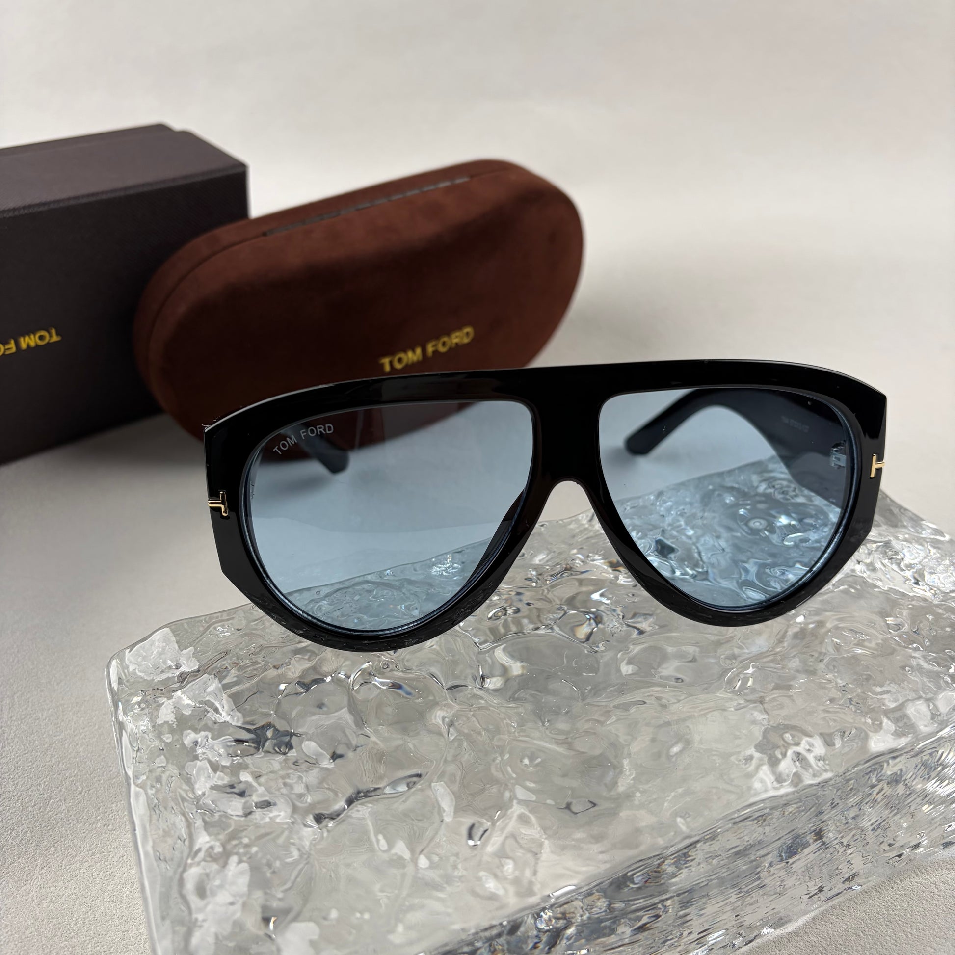 Black acetate oversized aviator sunglasses with blue tinted lenses resting on a clear ice-like block