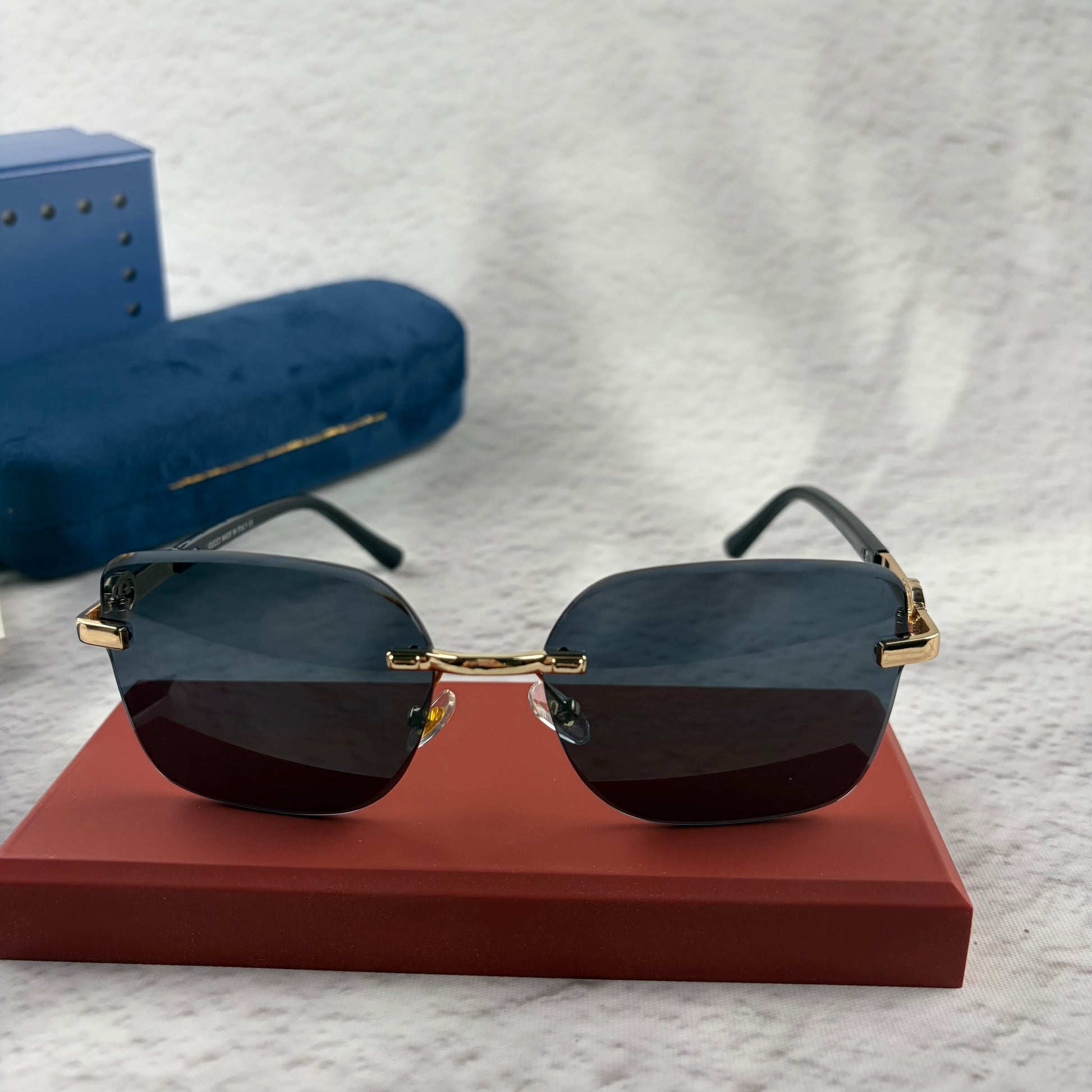Rimless rectangular sunglasses with gold-tone metal arms and dark lenses on a red display stand.