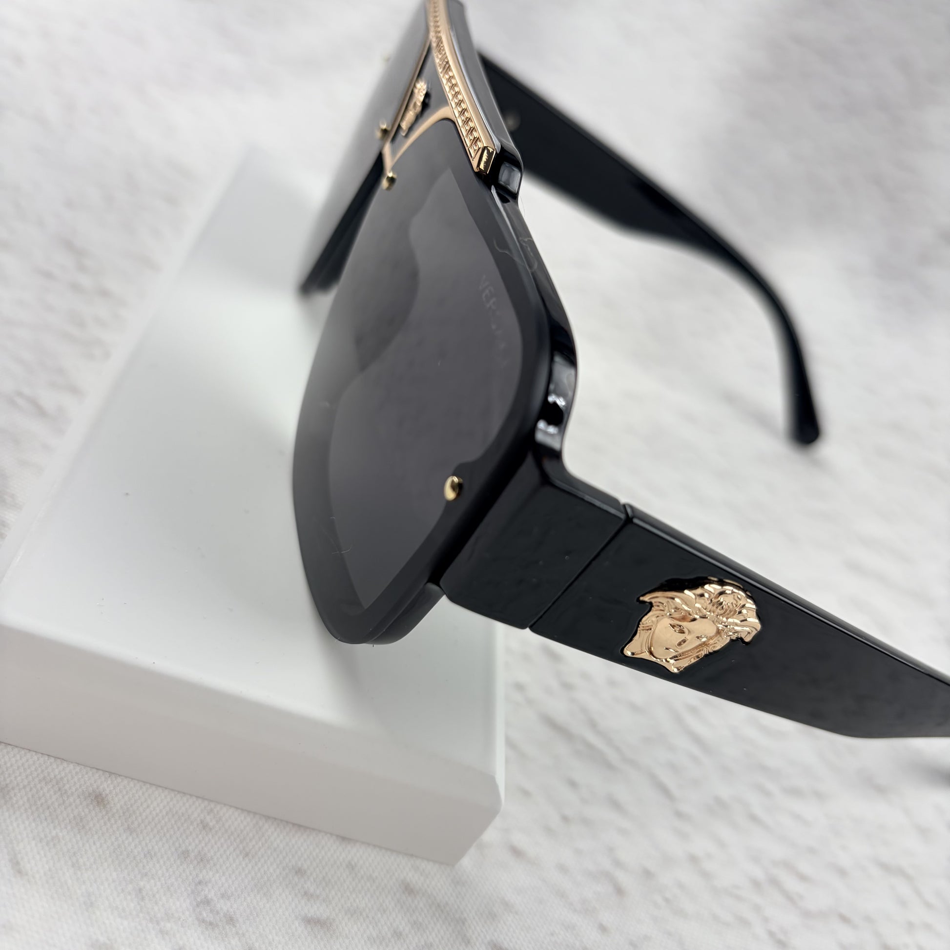 Oversized black flat-top sunglasses with a gold metal bridge and gold emblems on the temples.