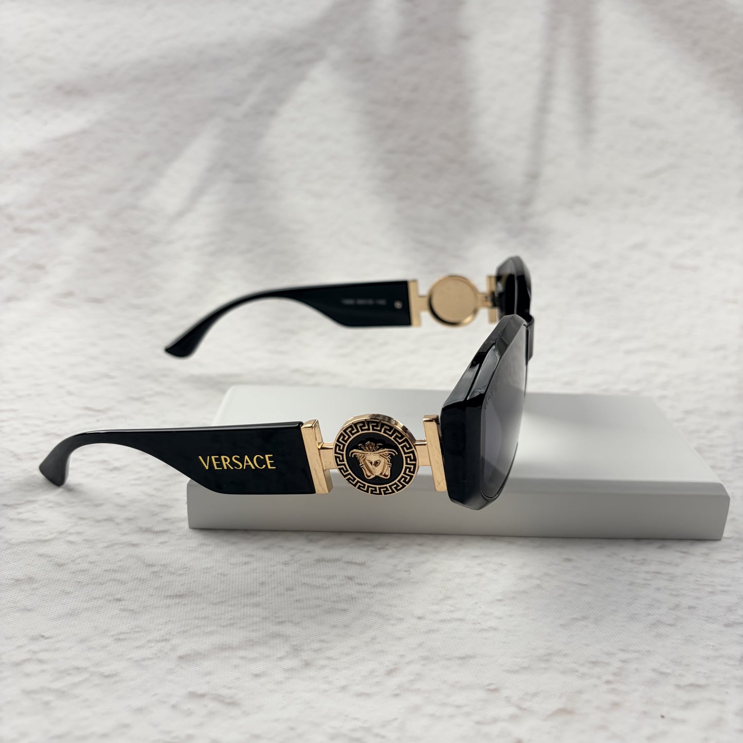 Black geometric sunglasses with gold side emblems resting on a white display block.