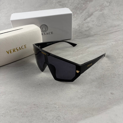 Oversized black shield sunglasses with gold metal accents on arms resting on a blue display box.