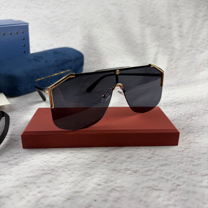 Oversized shield sunglasses with a gold metal frame and dark black lenses on a red display stand.