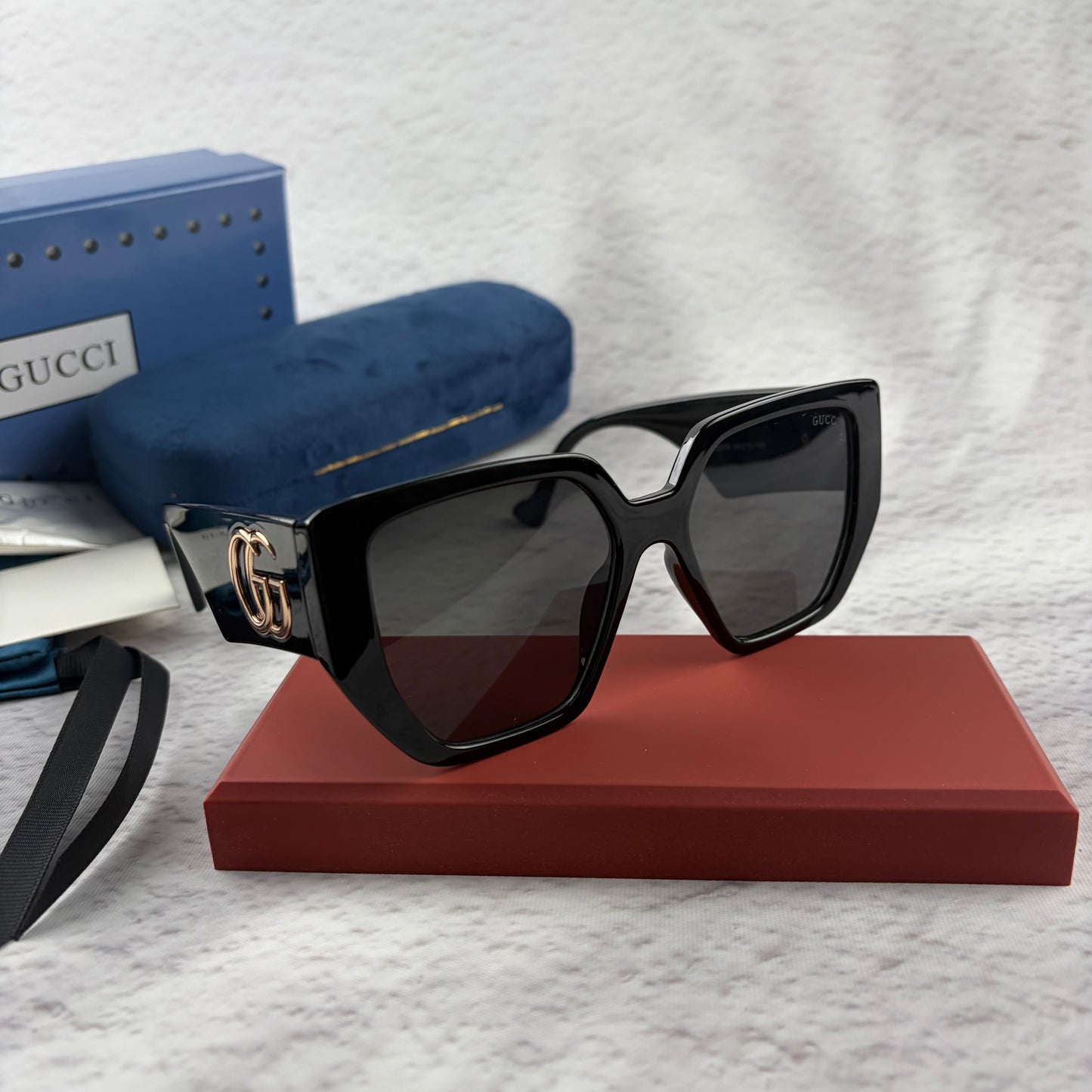 Oversized black square sunglasses with metallic temple detail, resting on a red display box.