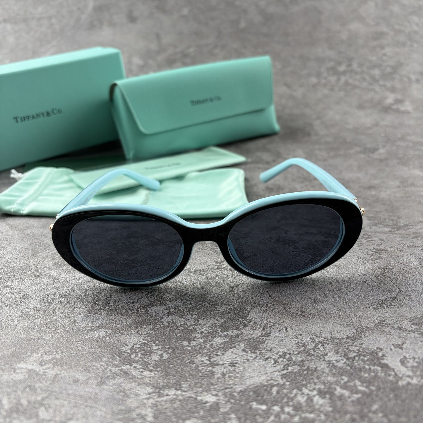 Elegant oval-frame sunglasses with contrasting color details and protective case on grey background.
