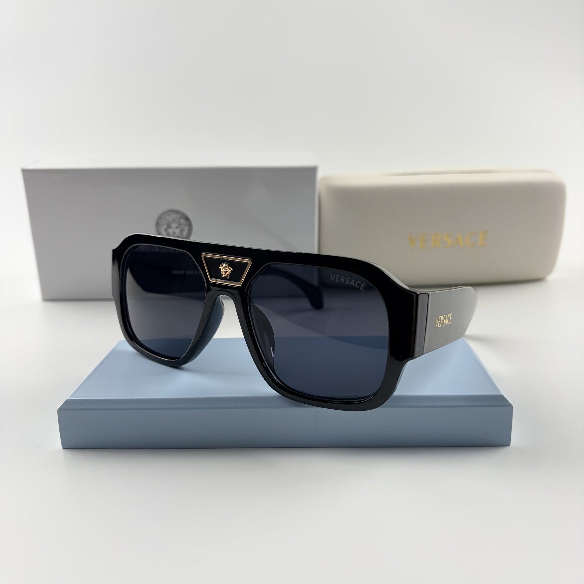 Black oversized aviator sunglasses with gold emblem on bridge, dark lenses, set on a blue stand.