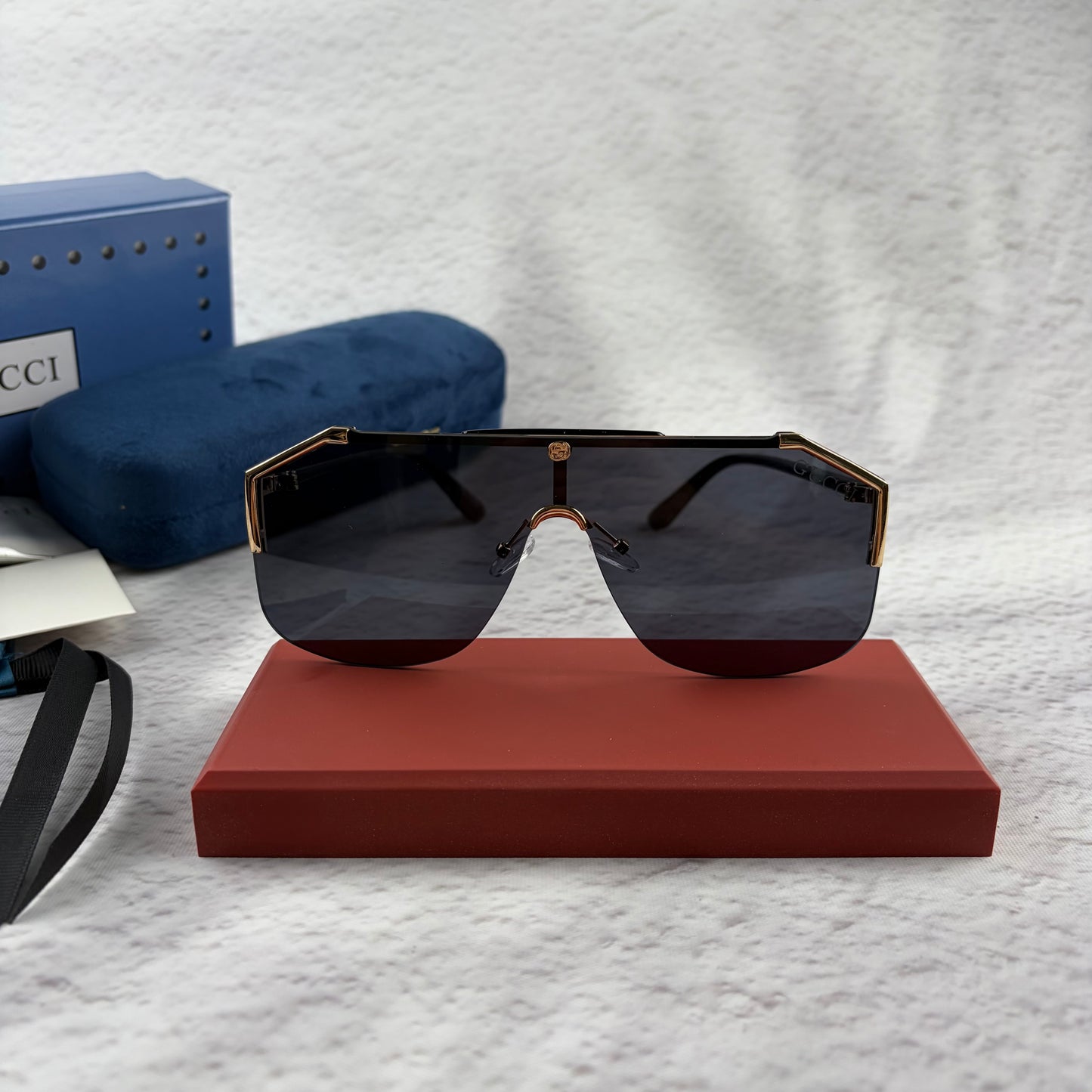 Oversized shield sunglasses with a gold metal frame and dark black lenses on a red display stand.