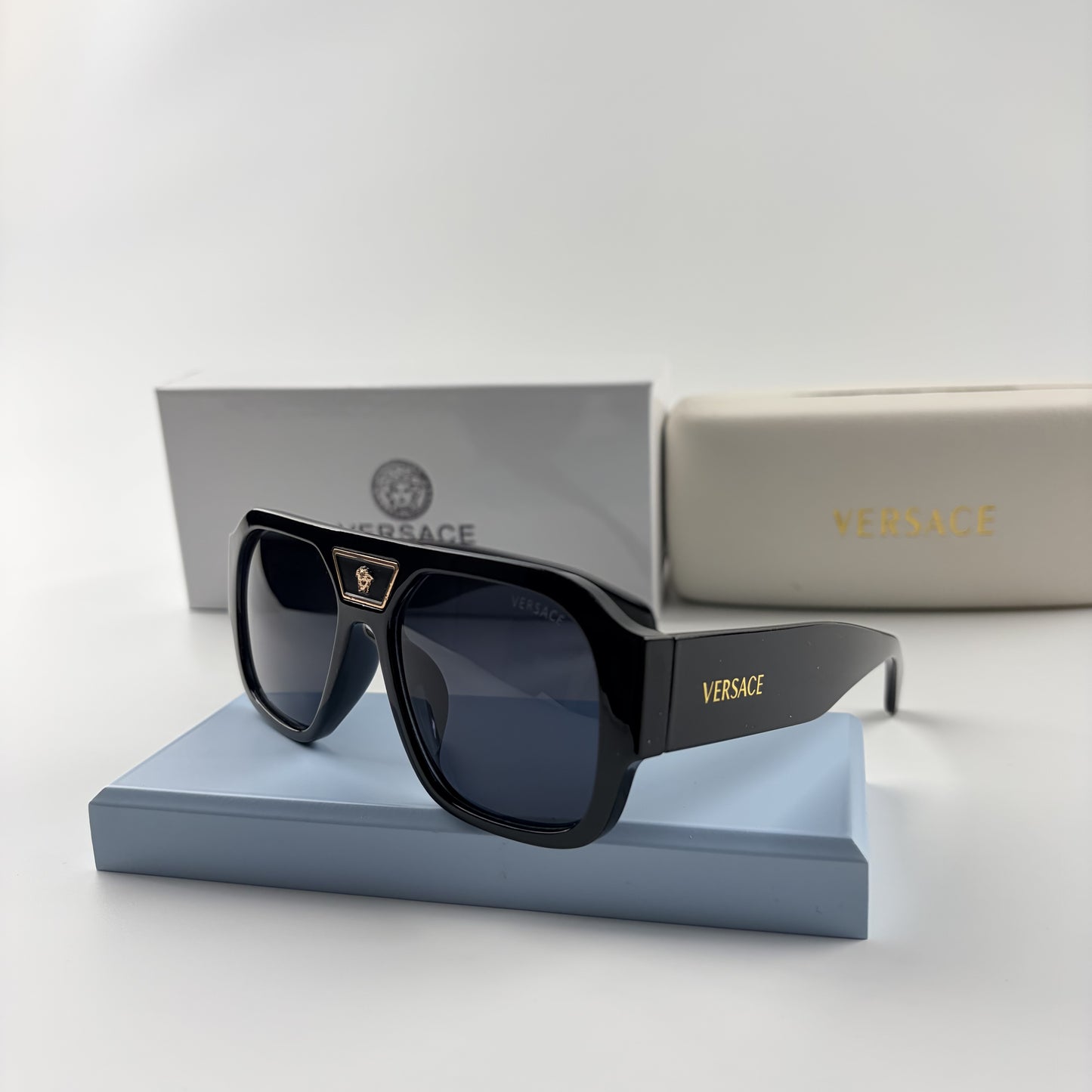 Black oversized aviator sunglasses with gold emblem on bridge, dark lenses, set on a blue stand.