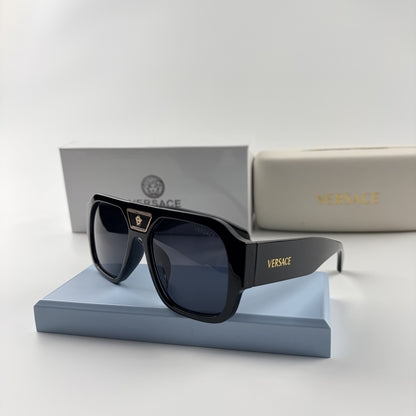 Black oversized aviator sunglasses with gold emblem on bridge, dark lenses, set on a blue stand.